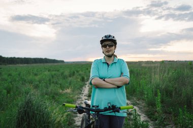 Kask takan bir kadın, kirli bir patika boyunca siyah bir dağ bisikleti sürüyor. Yol, arka planda evler ve ağaçların görülebildiği açık bir çayırdan geçiyor..