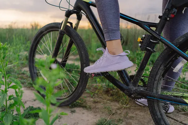 stock image A woman wearing white sneakers stands next to a mountain bike with a black tire on a dirt path. The bike is parked, and the woman is looking off-camera.