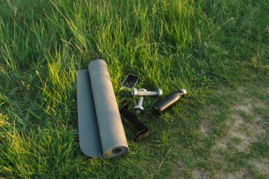 Eşyalar arasında bir yoga minderi, iki dambıl, bir siyah su şişesi ve bir akıllı telefon var. Bu resim açık havada egzersiz yapmanın kolaylığını tasvir ediyor, bir egzersiz için temel gereksinimlerin basitliğiyle..