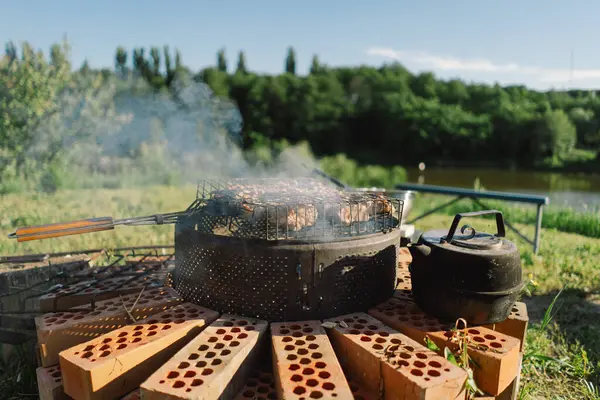 Stock image Barbecue set up on a sunny day near a river. The grill is filled with meat, and a kettle sits on a brick base beside it. Smoke rises from the grill, filling the air with the aroma of grilling food.