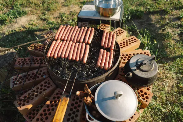 Stock image Barbecue set up on a sunny day near a river. The grill is filled with meat, and a kettle sits on a brick base beside it. Smoke rises from the grill, filling the air with the aroma of grilling food.