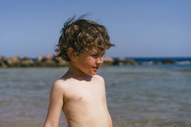 Kıvırcık kahverengi saçlı genç bir çocuk, arka planda kayalar ve okyanusla bir sahilde dururken kameraya gülümsüyor. Güneşli bir günde okyanusun sığ sularında koşan genç bir çocuk..