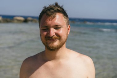 Kumsalda güneşli bir günde sığ suda dururken sakallı genç bir adam kameraya gülümsüyor. Yüzme şortu giyiyor ve rahat ve mutlu bir ifadesi var..
