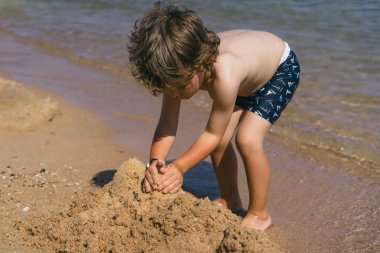 Kıvırcık kahverengi saçlı genç bir çocuk kumsalın kumlu sahilinde oturuyor, elleriyle kumdan kaleler inşa ediyor. Arkasından dalgalar yavaşça kıyıya vuruyor. Huzurlu bir manzara yaratıyor.