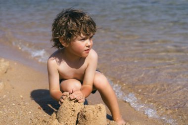 Kıvırcık kahverengi saçlı genç bir çocuk kumsalın kumlu sahilinde oturuyor, elleriyle kumdan kaleler inşa ediyor. Arkasından dalgalar yavaşça kıyıya vuruyor. Huzurlu bir manzara yaratıyor.