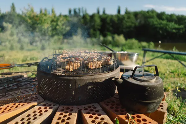 stock image Barbecue set up on a sunny day near a river. The grill is filled with meat, and a kettle sits on a brick base beside it. Smoke rises from the grill, filling the air with the aroma of grilling food.