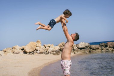 Bir baba oğluyla Sunny Beach 'te oynar. Oğlu okyanusa bakarken gülümsüyor. Baba sığ sularda duruyor, arka planda kayalar var..