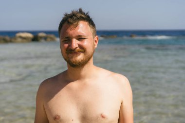 Kumsalda güneşli bir günde sığ suda dururken sakallı genç bir adam kameraya gülümsüyor. Yüzme şortu giyiyor ve rahat ve mutlu bir ifadesi var..