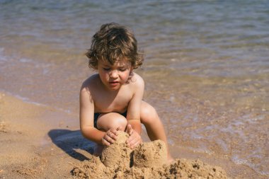 Kıvırcık kahverengi saçlı genç bir çocuk kumsalın kumlu sahilinde oturuyor, elleriyle kumdan kaleler inşa ediyor. Arkasından dalgalar yavaşça kıyıya vuruyor. Huzurlu bir manzara yaratıyor.
