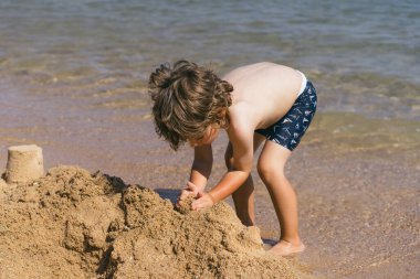 Kıvırcık kahverengi saçlı genç bir çocuk kumsalın kumlu sahilinde oturuyor, elleriyle kumdan kaleler inşa ediyor. Arkasından dalgalar yavaşça kıyıya vuruyor. Huzurlu bir manzara yaratıyor.