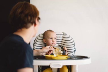 Bir kız bebek yüksek bir sandalyede oturuyor ve annesi tarafından besleniyor. Bebek sarı ve beyaz bir çiçek desenli gömlek giyiyor ve ellerini yüksek sandalyenin tepsisine koyuyor..