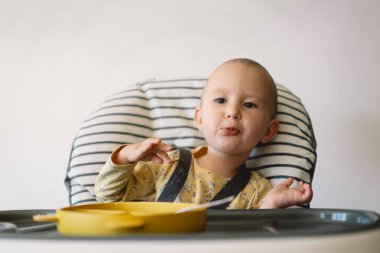 Bir kız bebek yüksek bir sandalyede oturuyor ve annesi tarafından besleniyor. Bebek sarı ve beyaz bir çiçek desenli gömlek giyiyor ve ellerini yüksek sandalyenin tepsisine koyuyor..