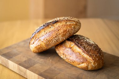 İki somun ekşi ekmek, taze pişmiş ve hala sıcak, ahşap bir yüzeye istiflenmiştir. Somunlar susam ve ayçiçeğiyle kaplıdır. Köy ve lezzet katar..