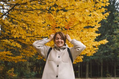 Bir kadın, canlı sonbahar yapraklarıyla bezenmiş ağaçlarla çevrili sakin bir ormanda yürüyor. Elinde taze toplanmış yapraklarla dolu bir sepet tutuyor. Mevsimin ılık renklerini sergiliyor.