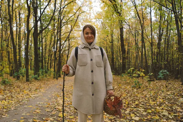 stock image A woman walks through a tranquil forest, surrounded by trees adorned with vibrant autumn foliage. She holds a basket filled with freshly gathered leaves, showcasing the warm colors of the season