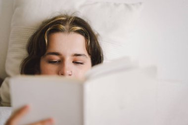 Bir kadın, elinde bir kupayla kanepede rahatlar ve sakin, davetkar bir atmosferle çevrili bir şekilde okumasına dalmıştır..