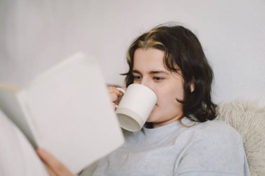 Bir kadın, elinde bir kupayla kanepede rahatlar ve sakin, davetkar bir atmosferle çevrili bir şekilde okumasına dalmıştır..