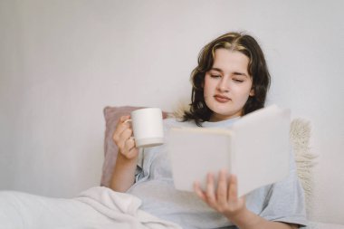 Bir kadın, elinde bir kupayla kanepede rahatlar ve sakin, davetkar bir atmosferle çevrili bir şekilde okumasına dalmıştır..