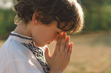 Çocuk, geleneksel işlemeli bir gömlek giyip, dua pozu vererek ayakta duruyor, ağaçların arasından süzülen yumuşak güneş ışığıyla sakin bir açık hava ortamının ortasında..