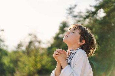 Çocuk, geleneksel işlemeli bir gömlek giyip, dua pozu vererek ayakta duruyor, ağaçların arasından süzülen yumuşak güneş ışığıyla sakin bir açık hava ortamının ortasında..