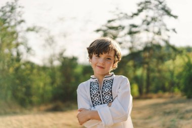 Genç bir çocuk, güneşli bir tarlada dikilirken özgüvenle gülümsüyor, öğleden sonra yemyeşil bir ortamda geleneksel işlemeli bir gömlek giyiyor.