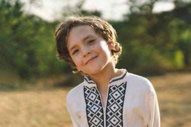 Genç bir çocuk, güneşli bir tarlada dikilirken özgüvenle gülümsüyor, öğleden sonra yemyeşil bir ortamda geleneksel işlemeli bir gömlek giyiyor.