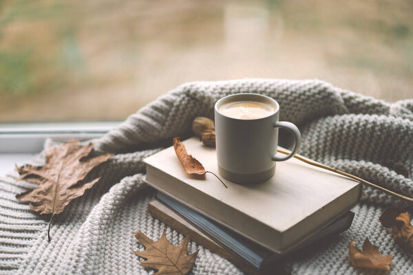 A warm cup of coffee rests on a stack of books, surrounded by soft blankets and autumn leaves, creating a tranquil atmosphere by the window.