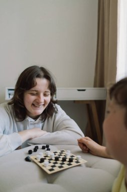 Genç kız ve oğlan oturma odasında masa oyunu oynuyorlar. Beyaz bir divanda masa oyunu oynuyorum. Onların her ikisi de oyuna odaklanmış. Çocuklar içeride dama tahtası oynuyor.