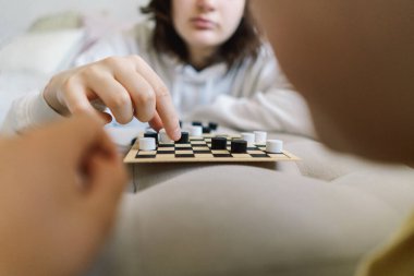 Genç kız ve oğlan oturma odasında masa oyunu oynuyorlar. Beyaz bir divanda masa oyunu oynuyorum. Onların her ikisi de oyuna odaklanmış. Çocuklar içeride dama tahtası oynuyor.