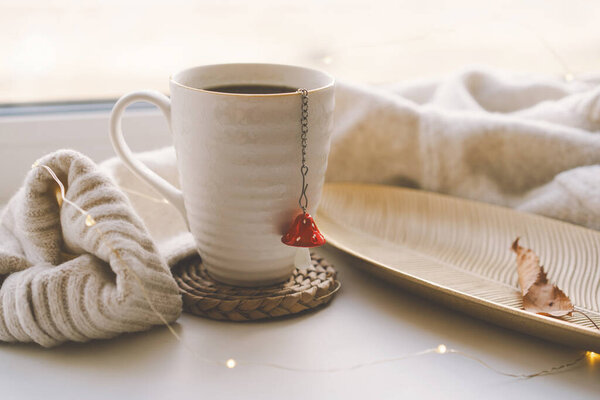 Парящая чашка чая лежит на сотканных горках рядом с маленькой декоративной тыквой, с мягкими вязаными одеялами и угощениями поблизости, освещенными теплым солнечным светом. Уютная осенняя концепция.