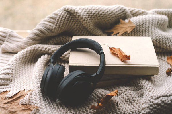 A pair of black headphones lies on an book, surrounded by autumn leaves and a knitted blanket, creating a warm and inviting atmosphere perfect for relaxation. Read, Rest. Cozy autumn or winter concept