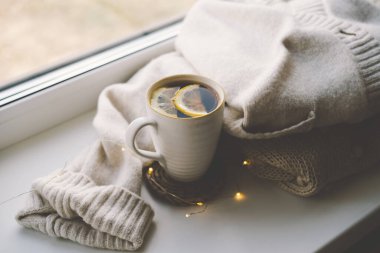 Limonlu dumanı tüten bir çay fincanı, yanında yumuşak örgü battaniyeler ve yakındaki sıcak güneş ışığıyla aydınlatılmış küçük bir balkabağının yanında örülmüş bir bardak altlığında duruyor. Samimi sonbahar konsepti.