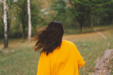 Parlak sarı bluzlu bir kadın, sonbaharda güneşin sıcaklığından ve doğanın güzelliğinden zevk alırken mutlu bir şekilde parkta yürür..