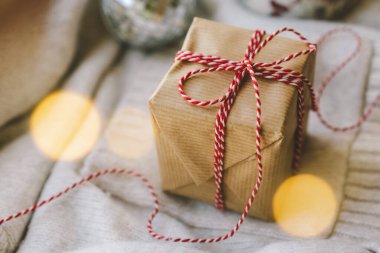 Kahverengi kağıt ve kırmızı kurdeleye sarılı küçük bir hediye yumuşak tatil ışıklarıyla çevrili yumuşak bir süvetere dayanır ve sıcak bir tatil atmosferi yaratır..