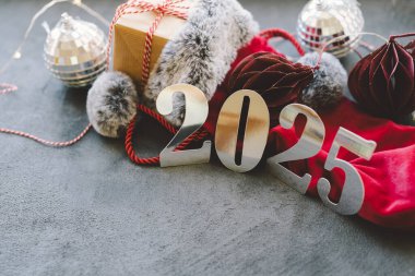 2025 yılı için parlak sayıların yanına ılık bir içecek konur, şenlikli süsler ve yumuşak dekorasyonlarla çevrili, rahat bir kış atmosferi yaratır. Mutlu Noeller Mutlu yıllar 2025