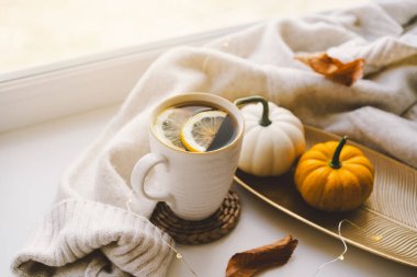 Limonlu dumanı tüten bir çay fincanı, yanında yumuşak örgü battaniyeler ve yakındaki sıcak güneş ışığıyla aydınlatılmış küçük bir balkabağının yanında örülmüş bir bardak altlığında duruyor. Samimi sonbahar konsepti.