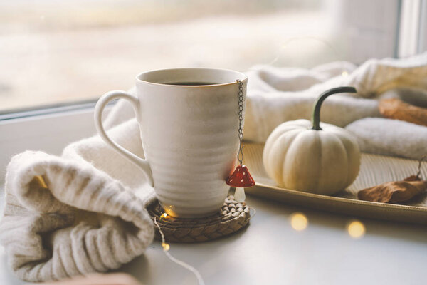 Парящая чашка чая лежит на сотканных горках рядом с маленькой декоративной тыквой, с мягкими вязаными одеялами и угощениями поблизости, освещенными теплым солнечным светом. Уютная осенняя концепция.