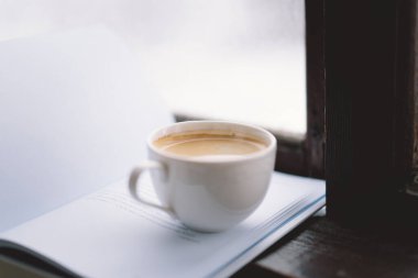 Sıcak bir fincan kahve pencerenin kenarında açık bir kitabın yanında durur, yanan bir mum yavaşça titreşir, karlı zemine karşı sakin bir atmosfer yaratır..