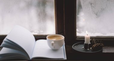 Sıcak bir fincan kahve pencerenin kenarında açık bir kitabın yanında durur, yanan bir mum yavaşça titreşir, karlı zemine karşı sakin bir atmosfer yaratır..