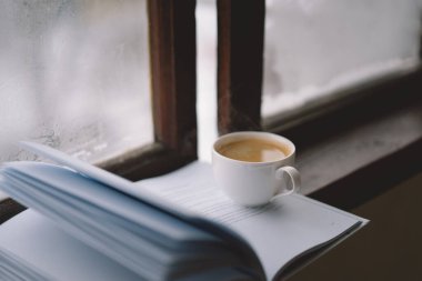 Sıcak bir fincan kahve pencerenin kenarında açık bir kitabın yanında durur, yanan bir mum yavaşça titreşir, karlı zemine karşı sakin bir atmosfer yaratır..