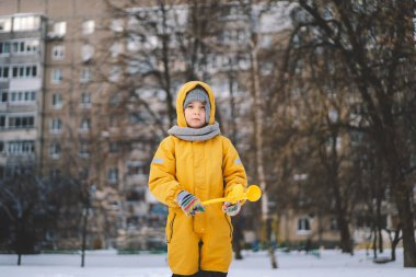 Soğuk bir kış günü, karlı bir arazide parlak sarı bir kış elbisesi giyen küçük bir çocuk bir kartopu makinesiyle oynuyor. Çocuk, sarı plastik bir kartopu makinesi kullanarak kartopu yapıyor.