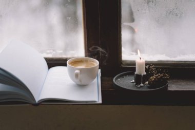 Sıcak bir fincan kahve pencerenin kenarında açık bir kitabın yanında durur, yanan bir mum yavaşça titreşir, karlı zemine karşı sakin bir atmosfer yaratır..