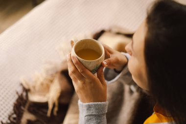 Bir kadın aydınlık, sakin bir odada rahatlar, kitabına odaklanırken kahvesini yudumlar. Pencerelerden gelen doğal ışık sıcak atmosferi güçlendirir..