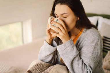Bir kadın aydınlık, sakin bir odada rahatlar, kitabına odaklanırken kahvesini yudumlar. Pencerelerden gelen doğal ışık sıcak atmosferi güçlendirir..