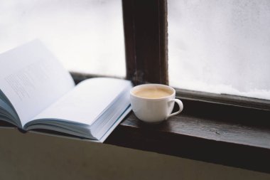 Sıcak bir fincan kahve pencerenin kenarında açık bir kitabın yanında durur, yanan bir mum yavaşça titreşir, karlı zemine karşı sakin bir atmosfer yaratır..