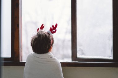 A young child wearing reindeer antlers sits wrapped in a cozy blanket, looking out a snow-covered window, evoking holiday spirit and warmth on a chilly day. clipart