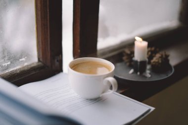 Sıcak bir fincan kahve pencerenin kenarında açık bir kitabın yanında durur, yanan bir mum yavaşça titreşir, karlı zemine karşı sakin bir atmosfer yaratır..