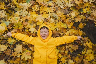Sarı yağmurluk giyen bir çocuk altın sonbahar yapraklarının arasında mutlu bir şekilde uzanır, gülümser ve serin sonbahar havasının tadını çıkarır. Yapraklar canlı bir zemin oluşturuyor, mevsimin güzelliğini gözler önüne seriyor.
