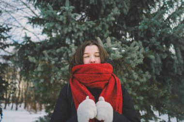 Kırmızı eşarplı gülümseyen bir kadın, parlak mavi gökyüzünün altındaki sakin bir ormanda karlı bir kış gününün tadını çıkarıyor. Noel ve Yeni Yıl Kutlama Konsepti.