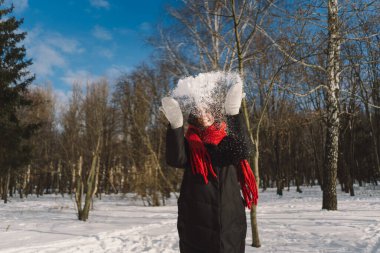 Kırmızı eşarplı gülümseyen bir kadın, parlak mavi gökyüzünün altındaki sakin bir ormanda karlı bir kış gününün tadını çıkarıyor. Noel ve Yeni Yıl Kutlama Konsepti.